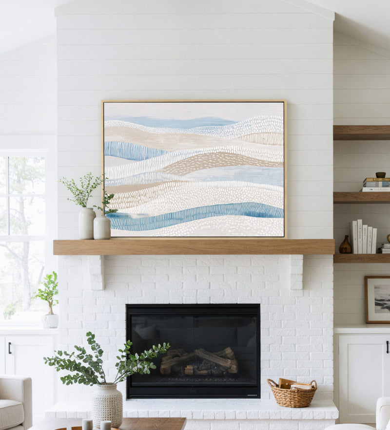 Large abstract coastal art print with muted blues and beige tones hanging above a white fireplace in a bright living room.