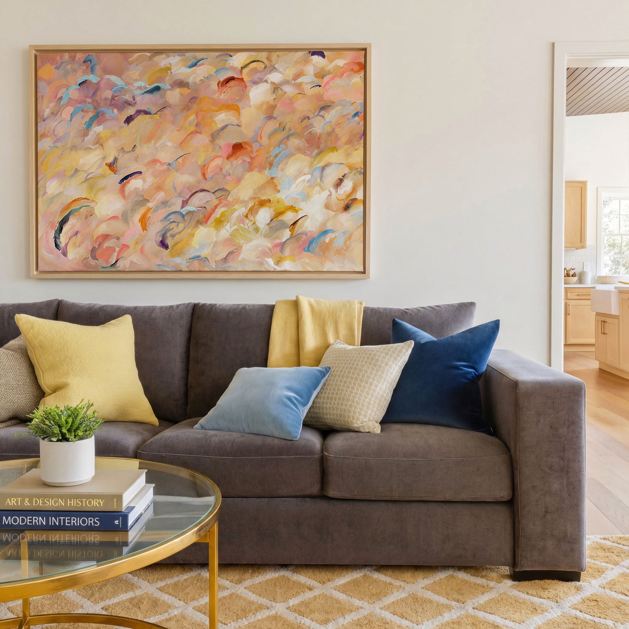 Warm abstract canvas print in natural wood floater frame above a grey sofa — cloud-like forms in peach, blush, warm yellow, coral, and soft blue with lavender and plum accents, styled in a bright contemporary living room with gold coffee table and mixed throw pillows
