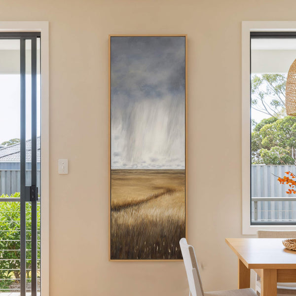This vertical canvas wall art features a moody landscape painting with a stormy gray sky and a golden field, framed in natural wood and hung on a light beige wall.
