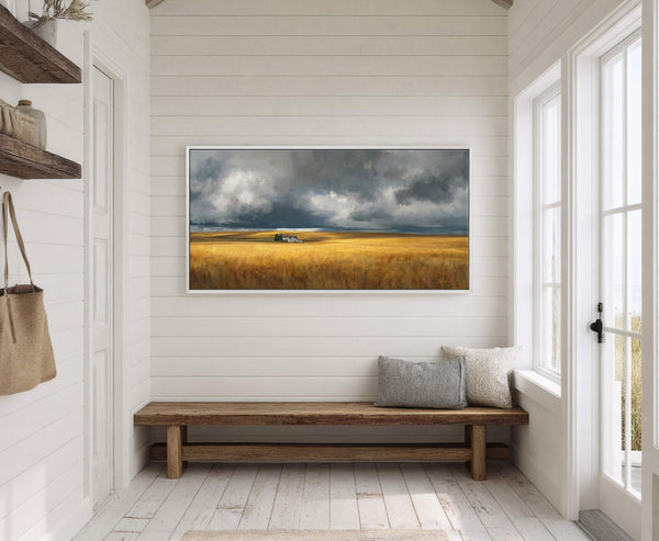 This horizontal canvas wall art features a rustic painting of a golden field under a stormy, dark sky with a small house. It is set against white shiplap walls, beside.