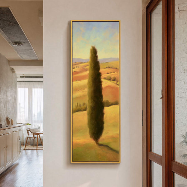 This vertical canvas wall art features a tall, dark green cypress tree in a rolling landscape with a warm, golden palette and impressionistic style, framed in gold, hanging on a.