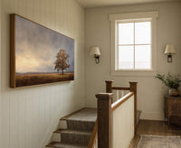 Horizontal canvas wall art depicts a solitary tree in a field under a cloudy sky, with a warm palette and realistic style. It is displayed in a hallway with white.