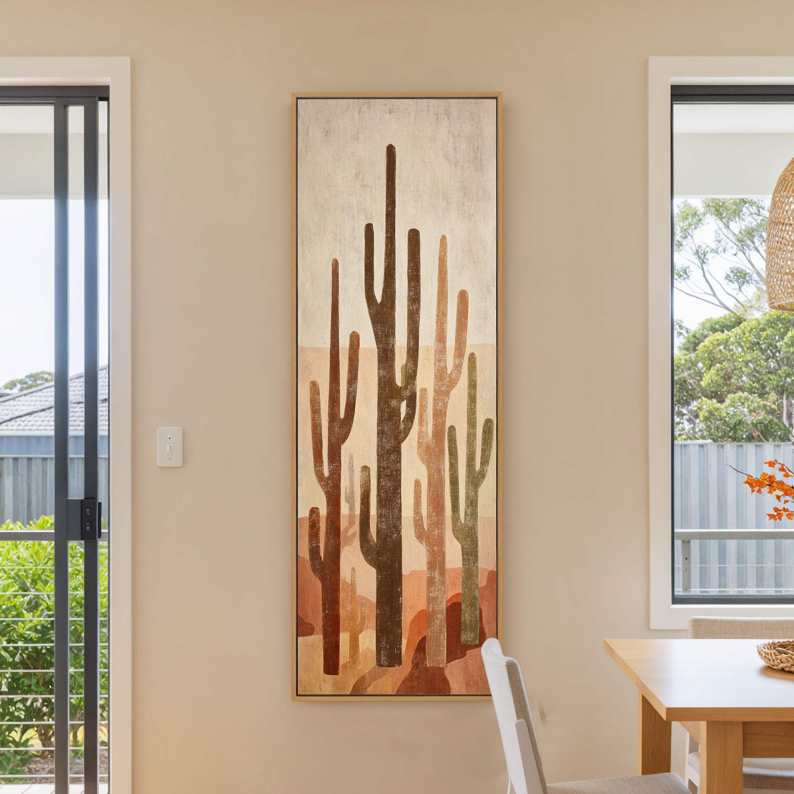 This vertical canvas print features a stylized desert landscape with saguaro cacti in muted browns and oranges, framed by a natural wood floater frame, displayed on a beige wall next.