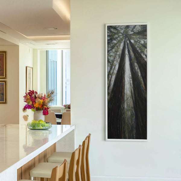 This vertical canvas wall art depicts a majestic view looking up at towering redwood trees with a lush green and brown palette, framed in white and displayed on a light-colored.