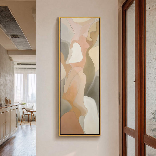 This vertical abstract art print features a soft, organic composition in a neutral palette of beige, blush, and gray, framed in gold and displayed on a beige wall beside a.