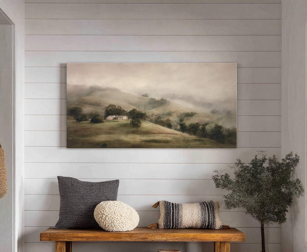 This horizontal canvas print features a misty landscape with rolling green hills and a distant house, rendered in soft, muted tones. It is displayed on a white shiplap wall in.