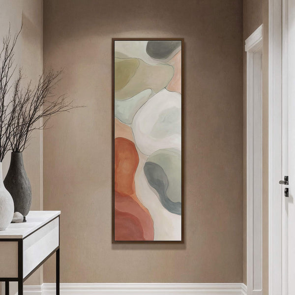 This vertical canvas wall art features an abstract organic design in a palette of muted greens, blush pink, terracotta, and cream, framed in dark wood, displayed on a tan wall.