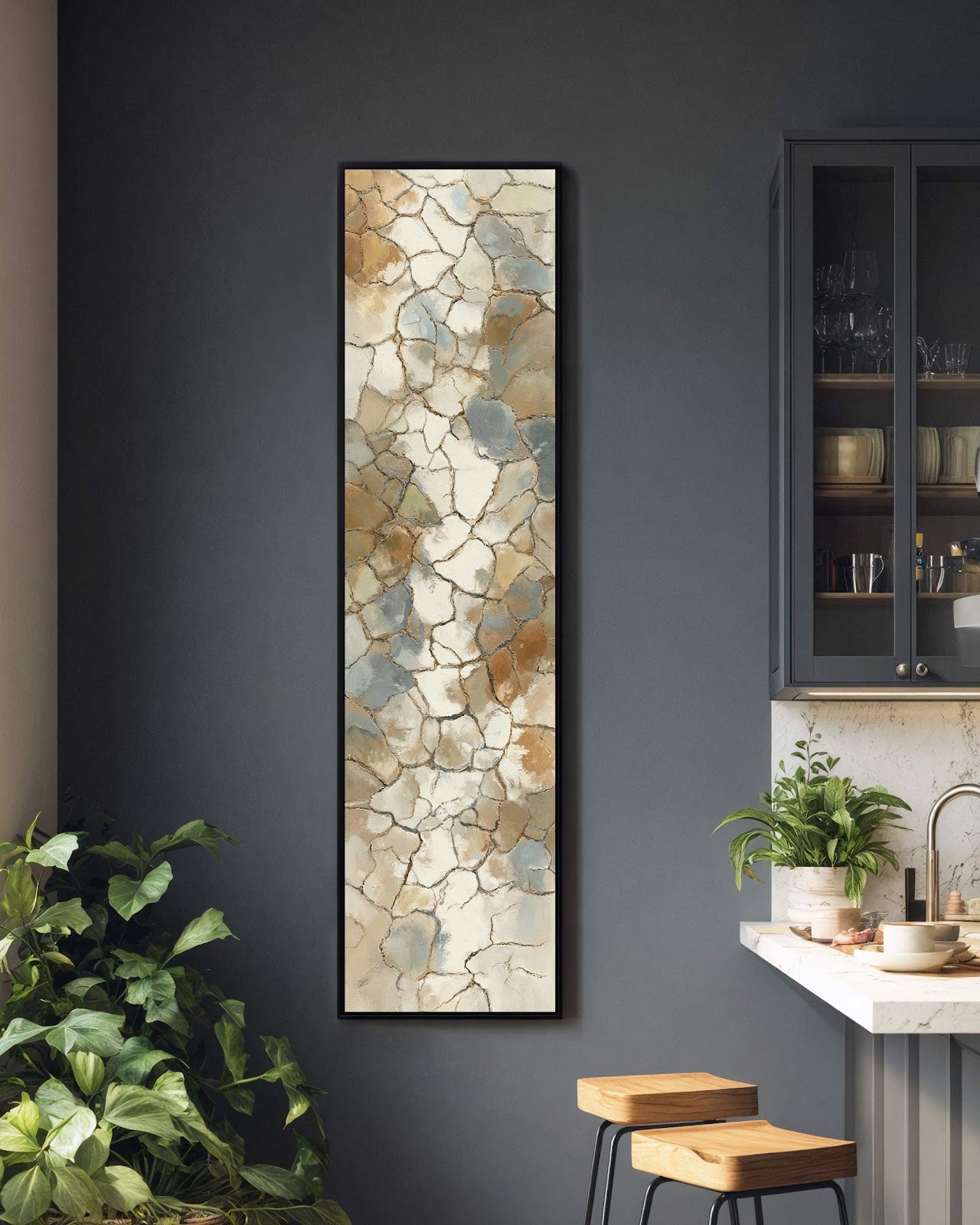This vertical art print displays a cracked abstract design in earthy beige and blue, showcased in a black floater frame on a dark gray kitchen wall beside a plant.