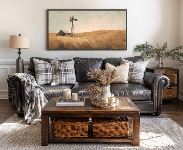 This horizontal canvas print depicts a rustic landscape with a windmill and barn in warm, muted tones and a painterly style, displayed above a dark leather sofa with plaid pillows.