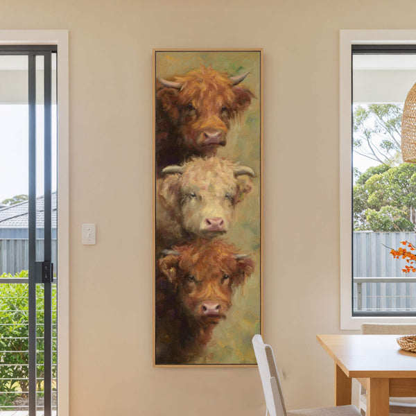 A vertical canvas print with a textured, impressionistic style depicts three Highland cows stacked vertically, featuring a warm brown and yellow palette, displayed on a neutral wall next to a.