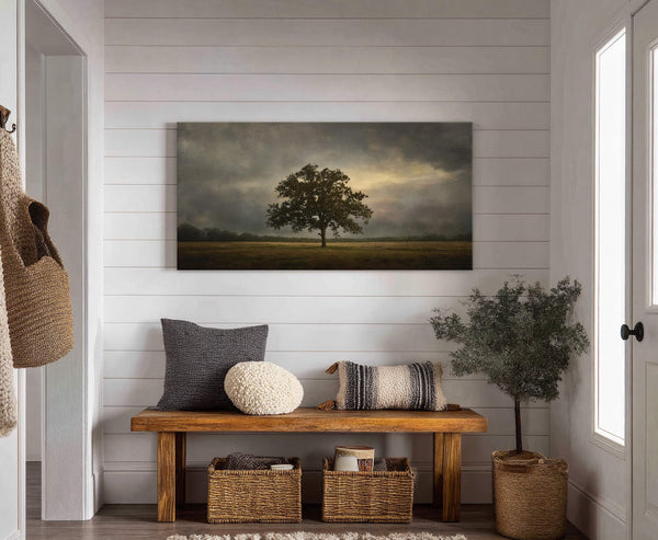 This horizontal canvas wall art features a painterly style with a muted palette of a lone tree against a dramatic, cloudy sky. It is displayed above a wooden bench with.