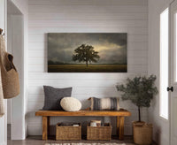 This horizontal canvas wall art features a painterly style with a muted palette of a lone tree against a dramatic, cloudy sky. It is displayed above a wooden bench with.
