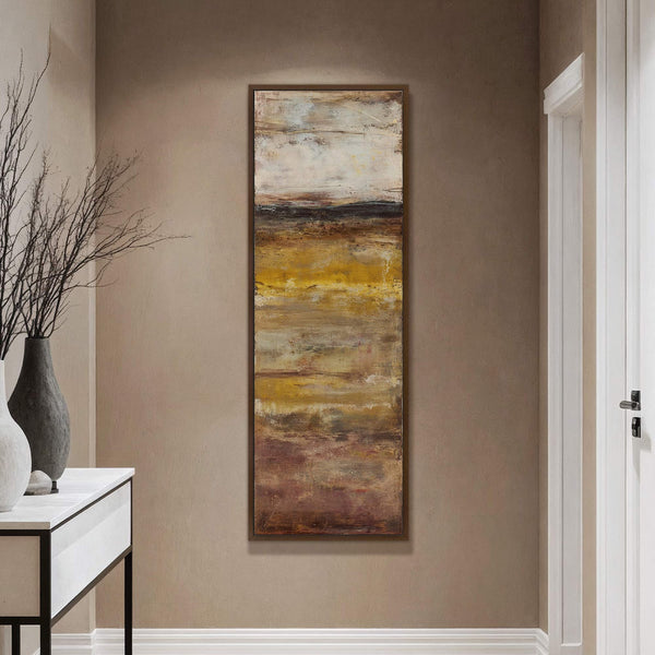 This vertical canvas wall art features an abstract landscape in earthy tones of brown, yellow, and white. It is framed in a dark brown wood and hangs on a tan.