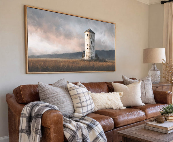 This horizontal canvas print showcases a moody, impressionistic landscape painting of a weathered white tower in a golden field under a dramatic, cloudy sky, displayed above a brown leather sofa.