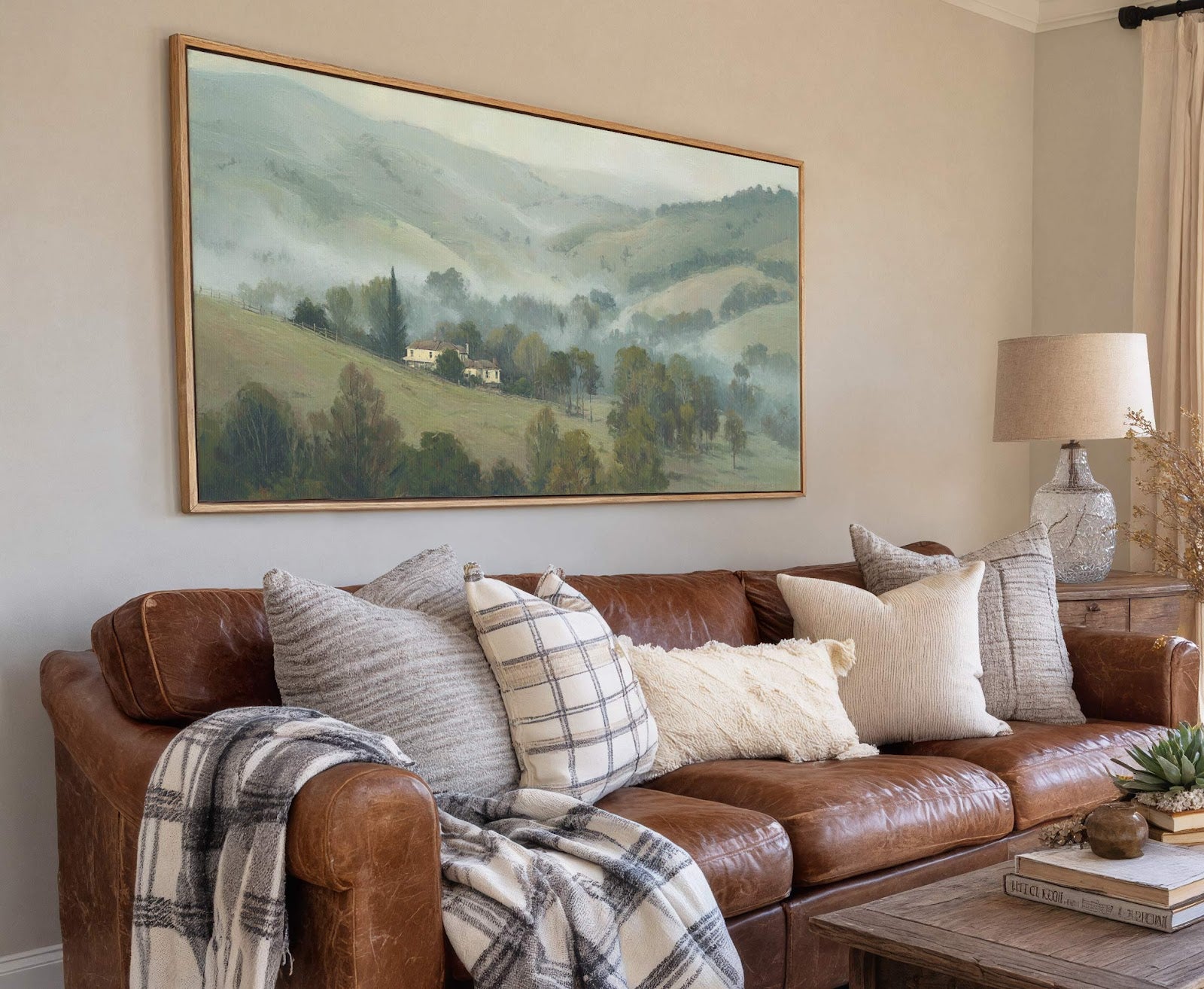 This horizontal canvas wall art features a misty, green landscape with rolling hills, trees, and a distant house, rendered in a soft, painterly style, displayed above a brown leather sofa.