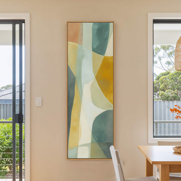 This vertical canvas print features an abstract organic style with a palette of muted greens, yellows, and oranges, displayed on a light tan wall next to a window and a.