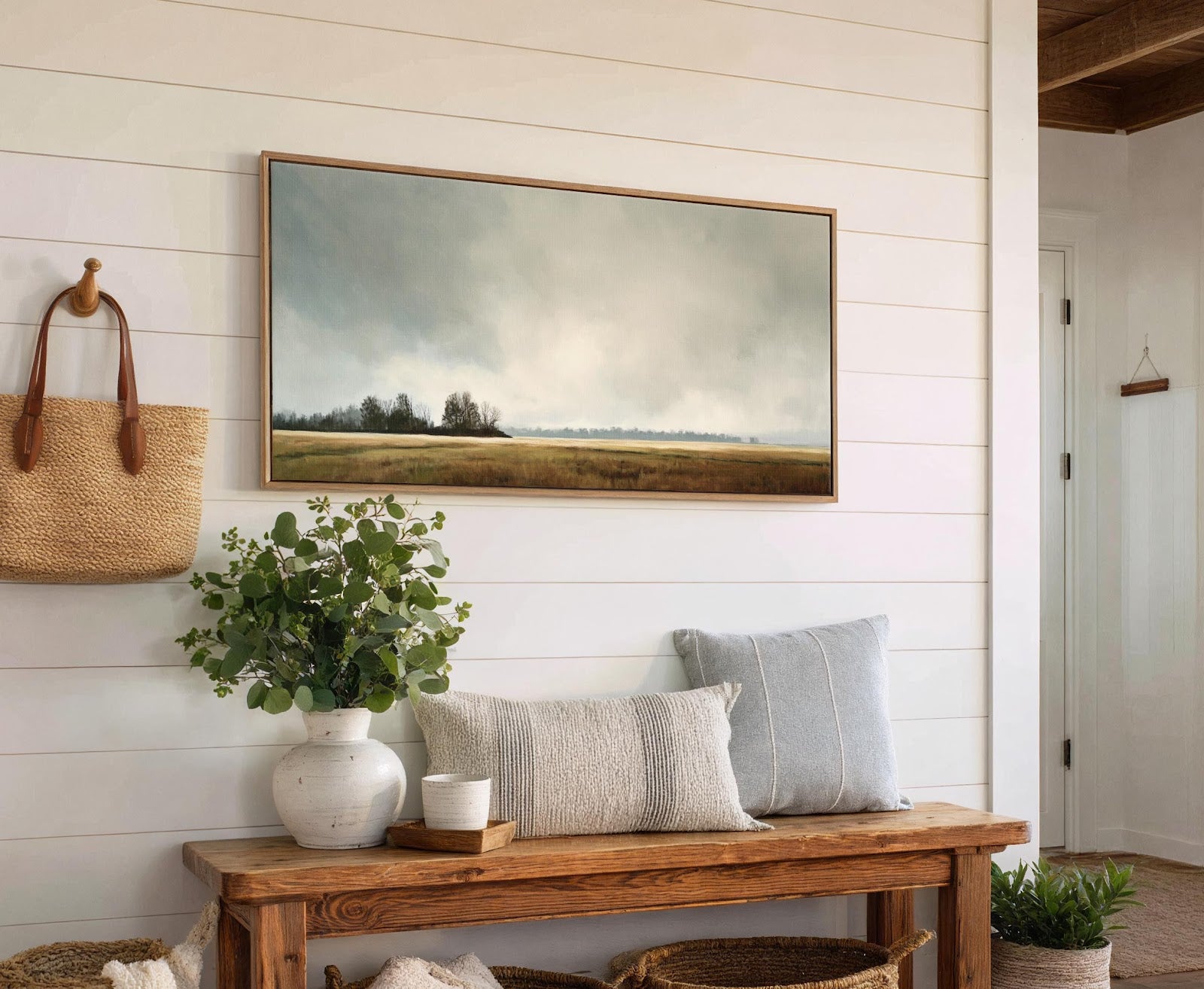 This horizontal canvas wall art depicts a moody landscape with a cloudy sky over a field and trees, framed in natural wood, hanging above a wooden bench on a white.