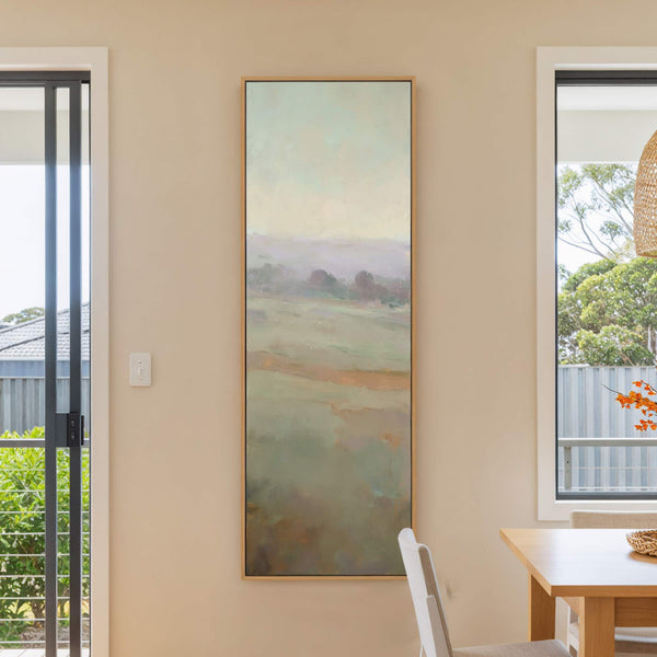 This vertical canvas print features an abstract landscape in muted greens and purples, with a soft pastel sky. It hangs on a beige wall next to a window and a.