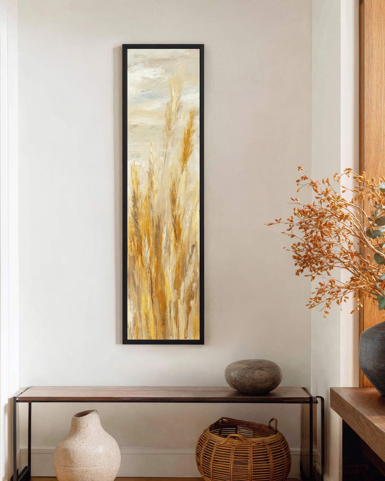 This vertical impressionistic art print features golden stalks in a black floater frame, displayed on a white wall above a minimalist wooden console table in a hallway.
