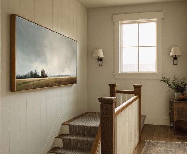 This horizontal canvas wall art showcases a muted landscape with trees against a cloudy sky, framed in natural wood, hanging on a paneled wall above stairs with a neutral rug.