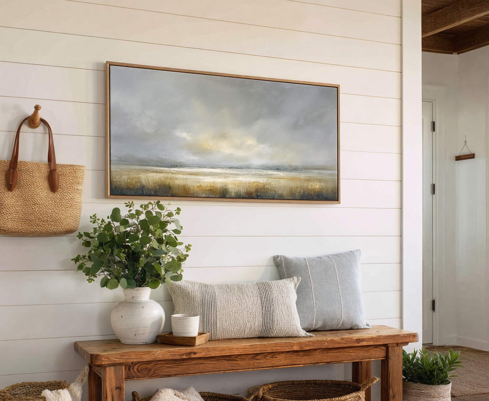 This horizontal canvas wall art features a moody landscape with a muted palette of grays, blues, and earthy tones, framed in natural wood, and hangs above a wooden bench with.