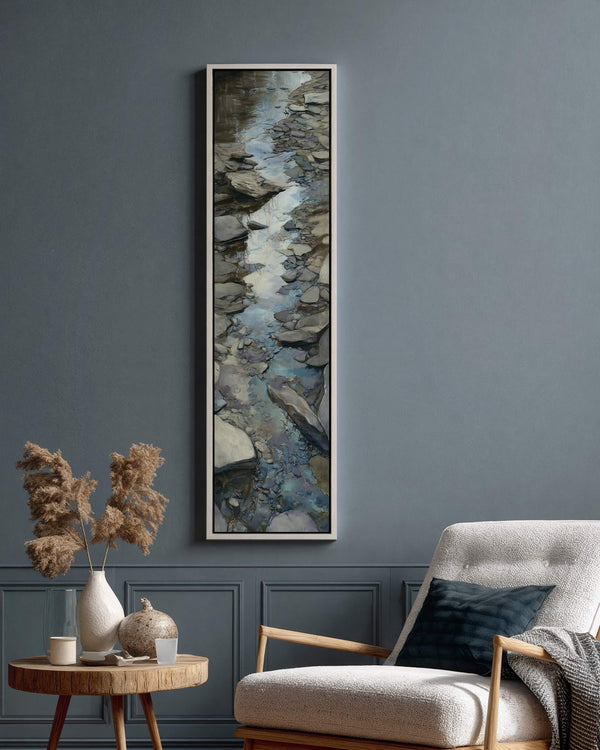 This vertical painterly canvas print of a rocky stream in blues and grays is displayed in a white floater frame on a blue wall beside a white armchair.