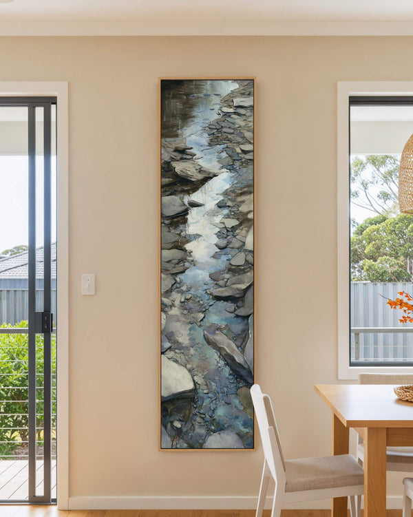 This vertical canvas art print features a painterly rocky stream in blue and gray, displayed in an oak floater frame on a beige dining room wall near a wooden table.
