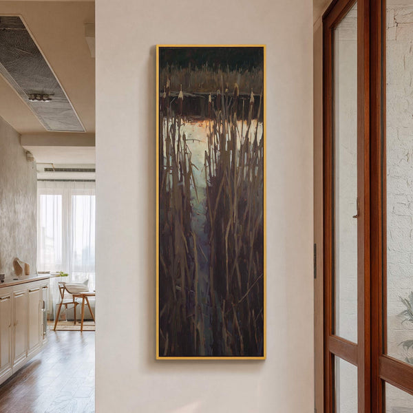 Vertical canvas wall art featuring an impressionistic painting of cattails against a body of water, with a dark, muted palette, framed in gold, displayed on a light beige wall near.