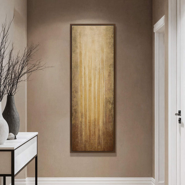 This vertical canvas wall art features an abstract expressionist style with a golden and brown palette, depicting vertical streaks of light. It is displayed in a room with taupe walls.