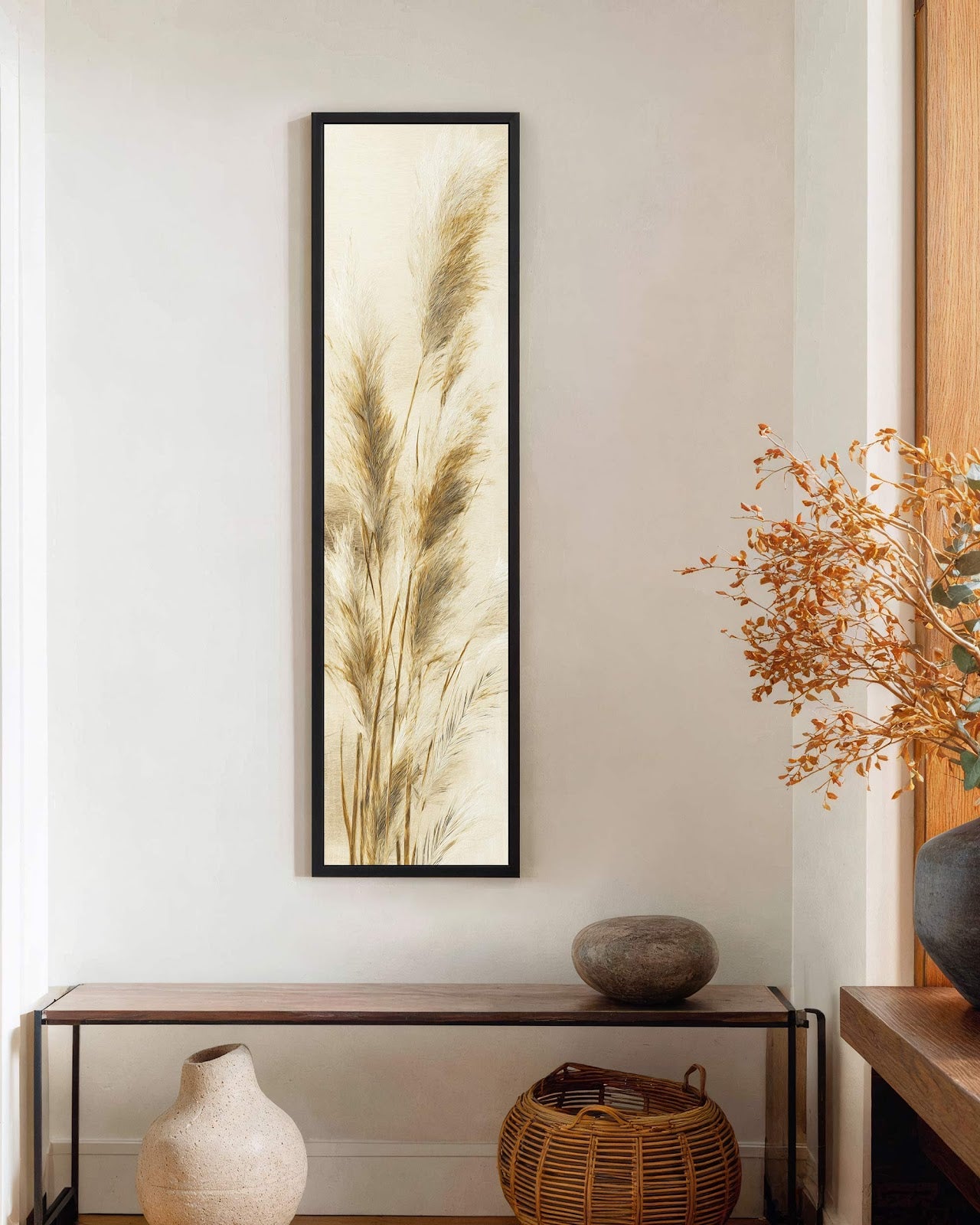 A vertical botanical painting print of beige pampas grass is displayed in a black floater frame above a wood console table on an off-white drywall living room wall.