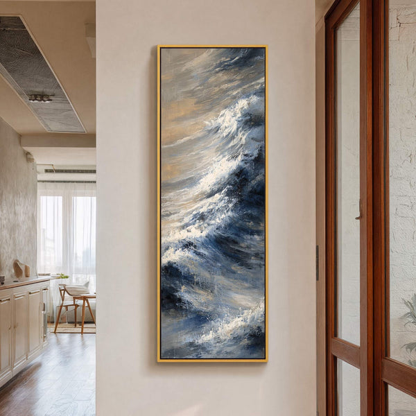 This vertical canvas wall art features an abstract depiction of ocean waves in a blue and beige palette, framed in gold, hanging on a neutral wall in a home interior.