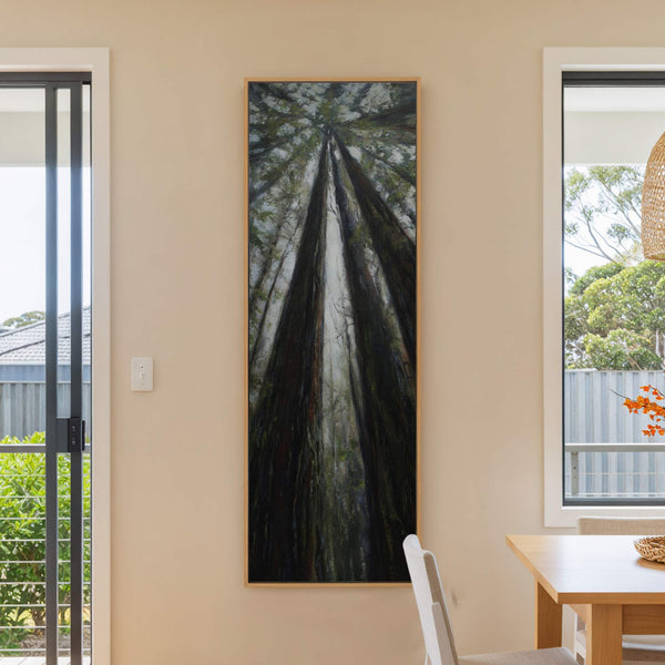 This vertical canvas wall art features a dark, moody impressionistic painting of redwood trees looking up towards the canopy, set against a neutral wall with a dining table visible.