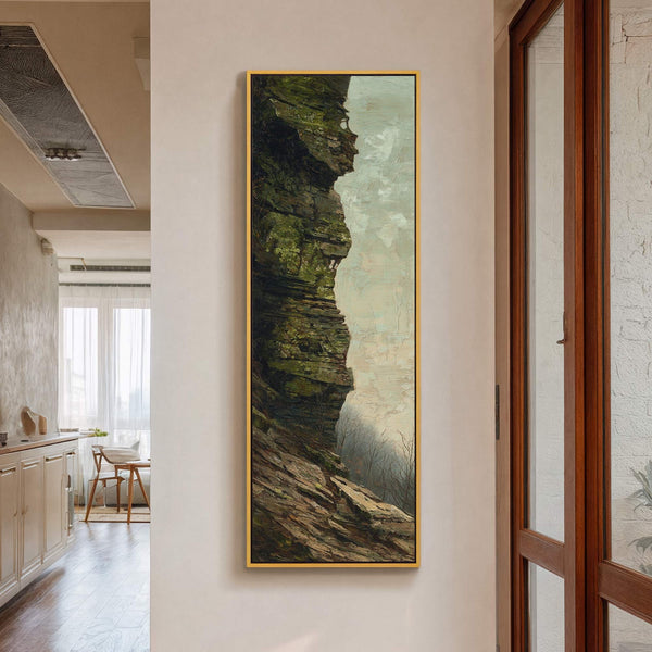 This vertical canvas wall art print features a rugged, moss-covered cliff face in muted greens and browns, against a hazy sky, framed in gold and hanging on a white wall.