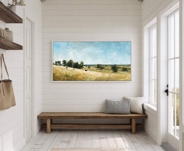 This horizontal canvas wall art depicts a pastoral landscape with cows grazing in a golden field under a soft blue sky, rendered in a painterly style. It hangs above a.
