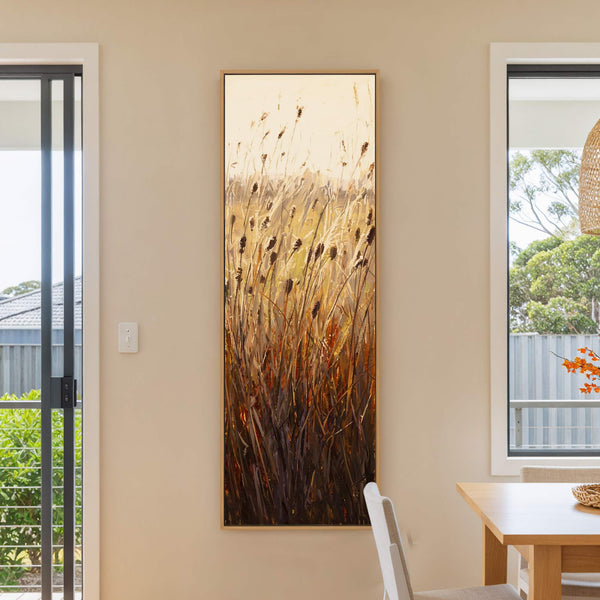 A vertical canvas print showcases a textured, impressionistic depiction of reeds in warm, earthy tones against a soft, beige wall. The artwork is framed in natural wood and hangs in.