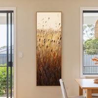 A vertical canvas print showcases a textured, impressionistic depiction of reeds in warm, earthy tones against a soft, beige wall. The artwork is framed in natural wood and hangs in.