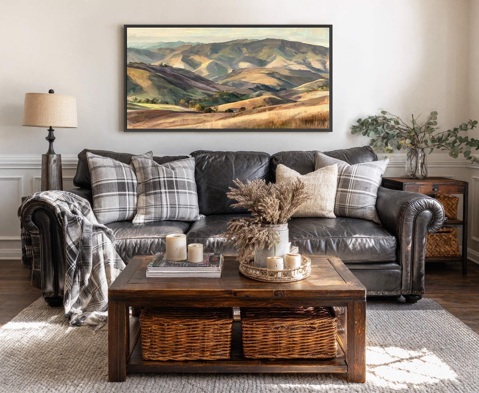This horizontal canvas wall art features a landscape painting with a palette of greens, browns, and blues, depicting rolling hills and fields, framed in black, displayed above a dark leather.