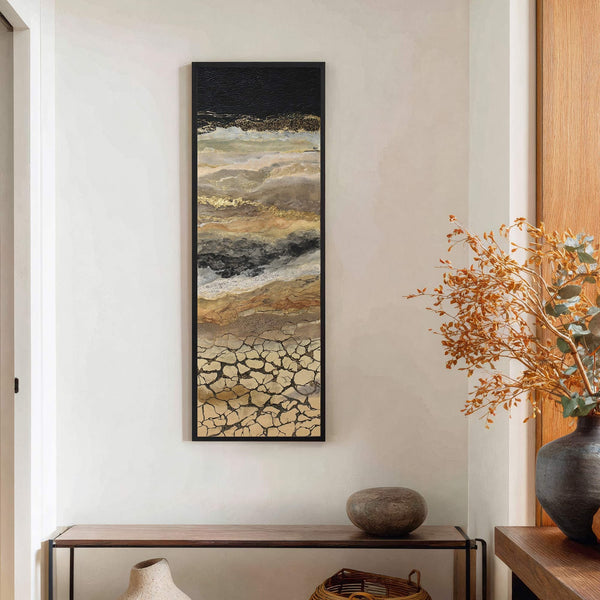 This vertical, abstract canvas wall art features a palette of black, gold, beige, and gray, evoking a cracked earth texture. It is displayed on a textured white wall above a.