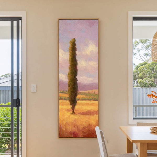 A vertical, impressionistic art print depicts a lone cypress tree in a sun-drenched field under a lavender sky, framed in natural wood and displayed on a neutral wall near a.