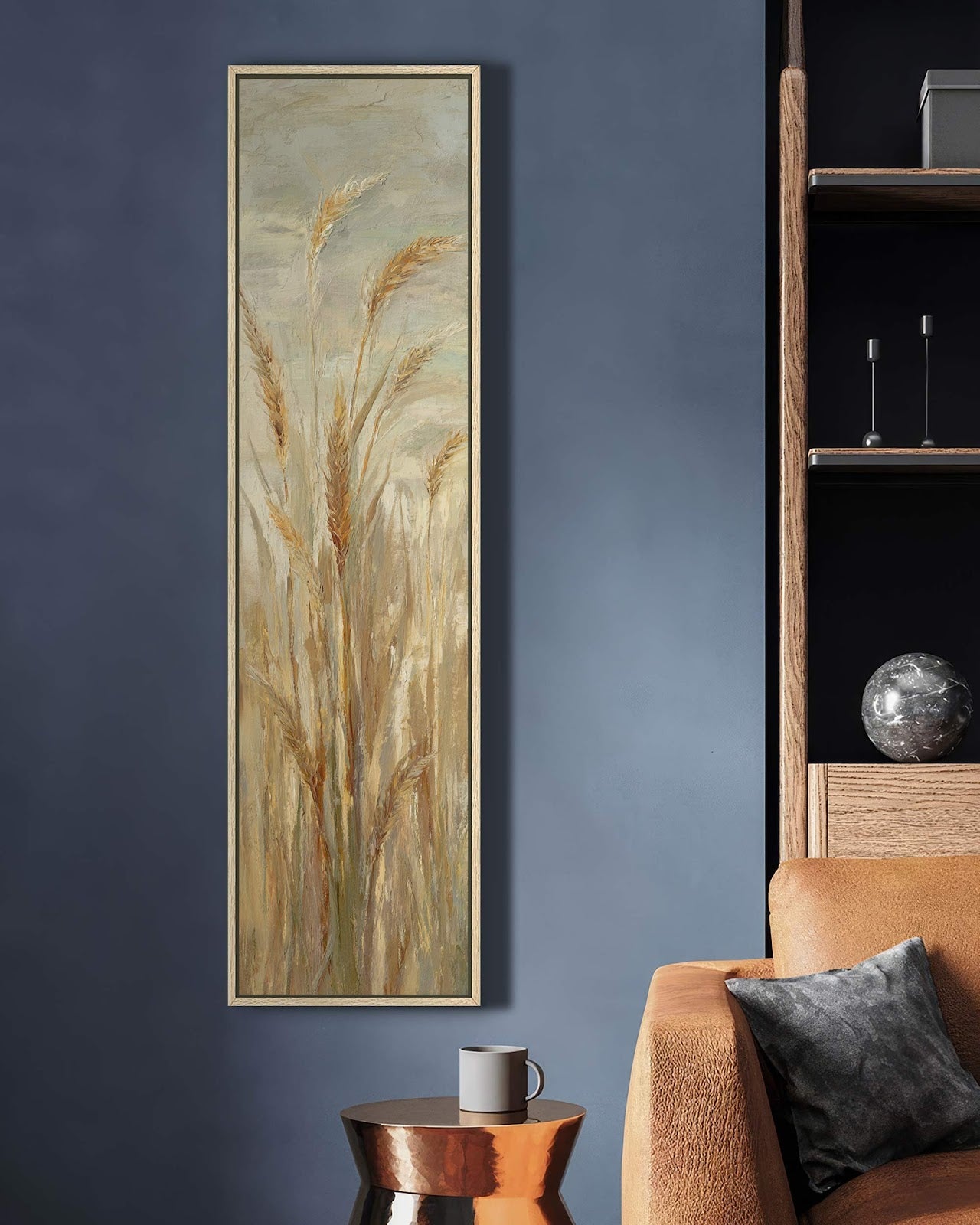 This vertical art print showcases painterly wheat stalks in earthy tones, presented in a natural oak floater frame on a dark blue living room wall beside a brown sofa.