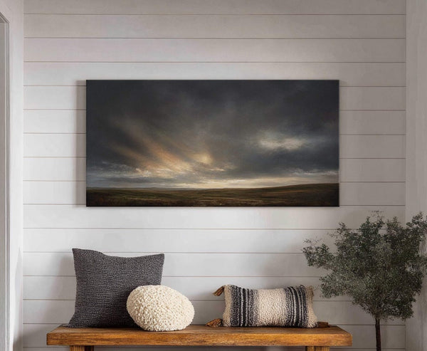 This horizontal canvas print depicts a dramatic, moody landscape with dark, cloudy skies and a golden sunset breaking through. It is displayed on a white paneled wall above a wooden.