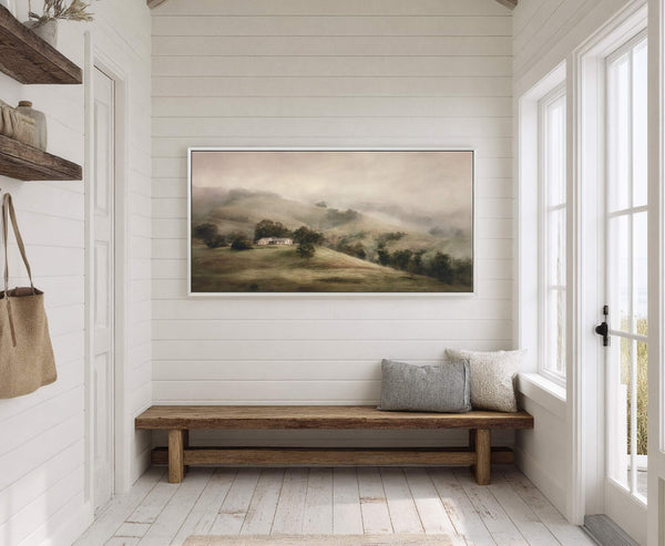 This landscape art print features a soft, muted palette of greens and browns depicting rolling hills shrouded in mist with a house on a hilltop, framed by a thin white.