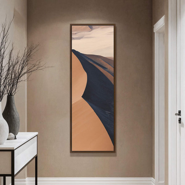 This vertical canvas print features an abstract desert landscape with a palette of warm oranges and deep blues, framed in a dark wood floater frame, displayed on a textured taupe.