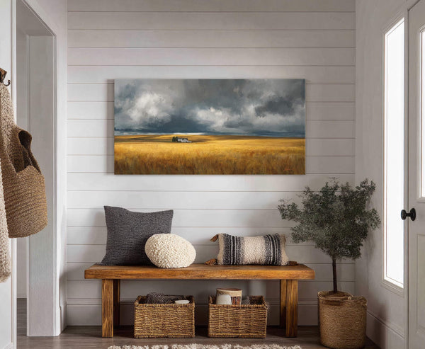 This horizontal canvas wall art features a dramatic landscape with a golden field and stormy clouds, displayed on a white shiplap wall above a wooden bench with decorative pillows and.