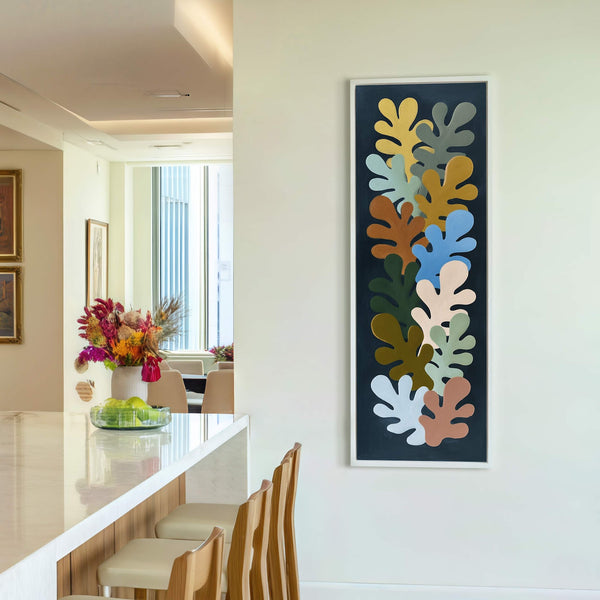 This vertical canvas wall art features a botanical motif in earthy tones and cool blues against a dark background, framed in white, and displayed on a white wall in-home wall.