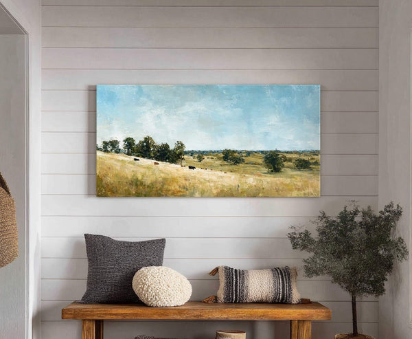 A horizontal canvas wall art print depicts a pastoral landscape with cows grazing on a golden field under a blue sky, displayed above a wooden bench with decorative pillows and.