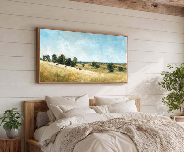 This horizontal canvas wall art features an impressionistic landscape of rolling hills and grazing cows under a bright blue sky, framed in natural wood, and displayed above a bed on.