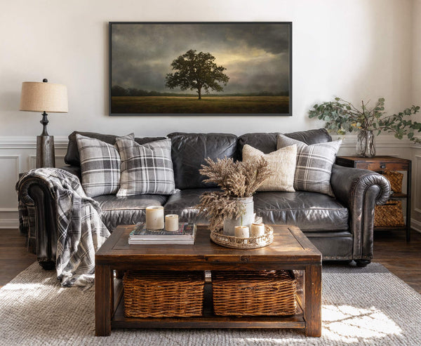 This canvas wall art features a dramatic landscape painting of a solitary oak tree against a cloudy, dramatic sky, framed in black and hung on a neutral wall in a.