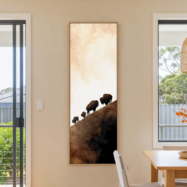 This vertical canvas wall art displays a silhouette of bison walking uphill against a muted watercolor sky, framed in natural wood, and hangs on a neutral wall in a room.