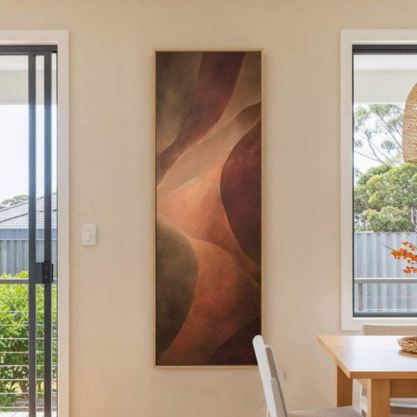 This vertical canvas wall art features an abstract design with a warm palette of browns, oranges, and reds, set against a neutral wall next to a window and a chair.
